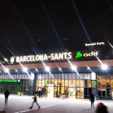 Noche en Barcelona Sants. Luces brillantes iluminan la fachada de la estación, con letreros de "Farmàcia" y "McDonald's".