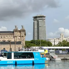 Barco turístico azul navega en el hafen Barcelona, con edificios históricos y modernos de fondo.