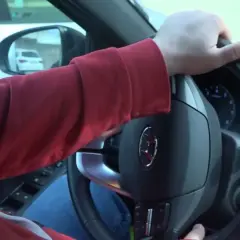 Mano girando el volante, se escucha un ruido al girar el volante. El conductor lleva una sudadera roja y va en un coche.
