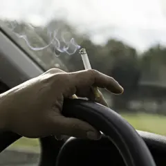 Mano sujetando un cigarrillo encendido sobre el volante. El humo se eleva mientras se puede fumar conduciendo.
