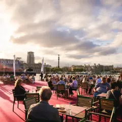 Gente disfrutando en terrazas al aire libre en el bsm moll de la fusta, con el mar y edificios de Barcelona de fondo.
