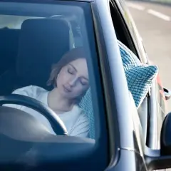 Mujer rubia duerme en el coche, apoyada en una almohada azul. Parece que se puede dormir en el coche cómodamente.