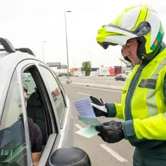 Agente de la Guardia Civil, con casco y chaleco reflectante, se acerca a un coche para entregar un documento. Parece que prescribe una multa.