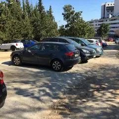 Aparcamiento del zoo de Barcelona con varios coches aparcados bajo árboles frondosos y un edificio moderno al fondo.
