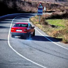 Coche rojo derrapando en curva, provocando subviraje y sobreviraje. Humo azul sale de las ruedas. Señal de curva peligrosa y límite de 60.