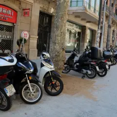 Un estacionamiento de motos T1 en una calle urbana. Varias motos y scooters están aparcados en la acera frente a edificios con tiendas y restaurantes.