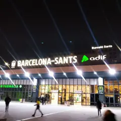 Noche en Barcelona Sants. Luces brillantes iluminan la fachada de la estación, con letreros de "Farmàcia" y "McDonald's".