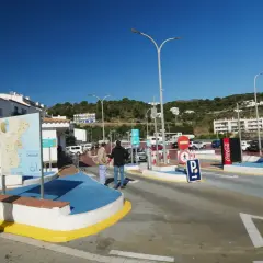 Entrada a parking gratuit Cadaqués. Una mujer rubia sonríe cerca de un mapa turístico. El cielo azul y las casas blancas dominan la escena.