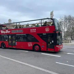 Autobús rojo de dos pisos "City Tour Barcelona" con turistas en la cubierta superior, ideal para el transporte en Barcelona para turistas.