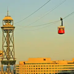 La cabina roja del montjuic seilbahn viaja sobre la ciudad, con una torre de transmisión y edificios modernos de fondo.