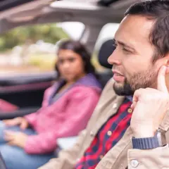 Hombre en coche toca su oreja, preocupado por un ruido al frenar a baja velocidad. Mujer sentada detrás, con expresión seria.