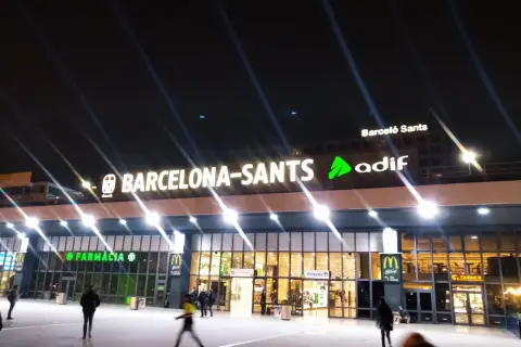 Noche en Barcelona Sants. Luces brillantes iluminan la fachada de la estación, con letreros de "Farmàcia" y "McDonald's".