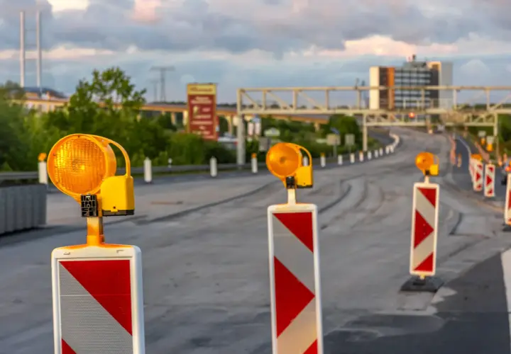 Una hilera de postes con luces naranjas intermitentes y marcas rojas y blancas, una señal de balizamiento, guía el tráfico en una carretera en construcción.