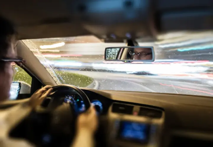 Conduciendo de noche bajo la lluvia, las luces de la carretera se difuminan. El conductor, con gafas, se refleja en el retrovisor, mostrando que es el ASR de un coche.