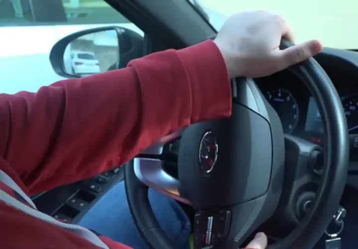 Mano girando el volante, se escucha un ruido al girar el volante. El conductor lleva una sudadera roja y va en un coche.