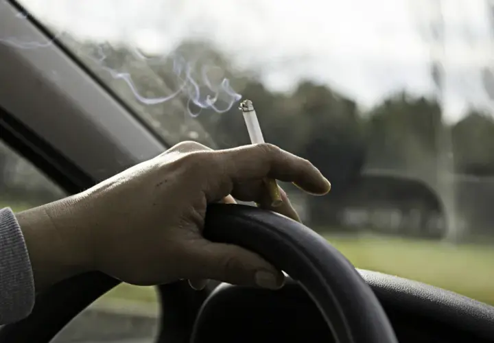 Mano sujetando un cigarrillo encendido sobre el volante. El humo se eleva mientras se puede fumar conduciendo.