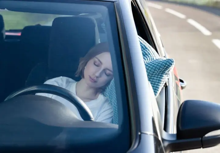 Mujer rubia duerme en el coche, apoyada en una almohada azul. Parece que se puede dormir en el coche cómodamente.