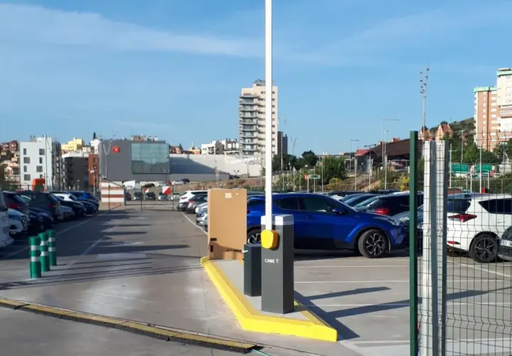Amplio parking BSM Vall d'Hebron con coches aparcados y barrera de acceso. Edificios modernos y colinas al fondo.
