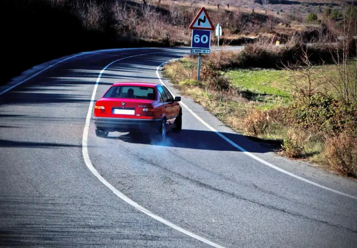 Coche rojo derrapando en curva, provocando subviraje y sobreviraje. Humo azul sale de las ruedas. Señal de curva peligrosa y límite de 60.