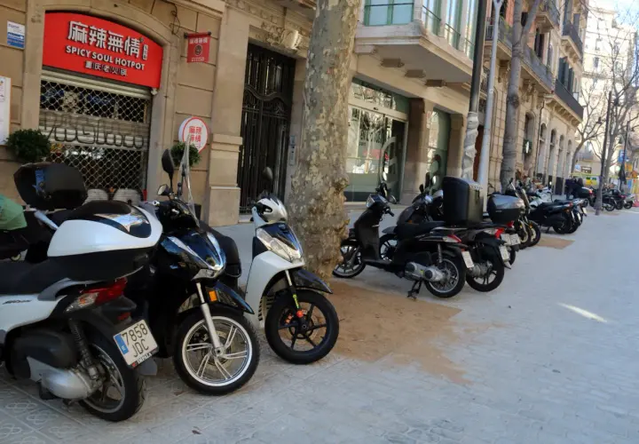 Un estacionamiento de motos T1 en una calle urbana. Varias motos y scooters están aparcados en la acera frente a edificios con tiendas y restaurantes.