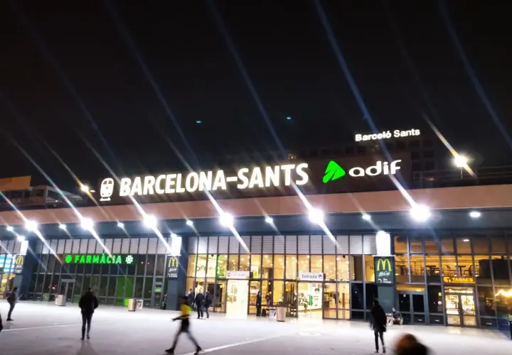 Noche en Barcelona Sants. Luces brillantes iluminan la fachada de la estación, con letreros de "Farmàcia" y "McDonald's".