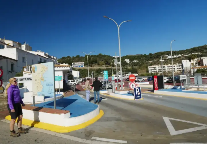 Entrada a parking gratuit Cadaqués. Una mujer rubia sonríe cerca de un mapa turístico. El cielo azul y las casas blancas dominan la escena.