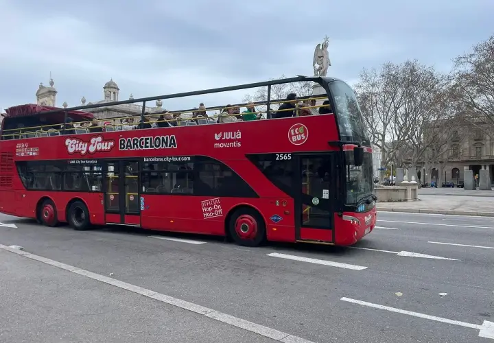 Autobús rojo de dos pisos "City Tour Barcelona" con turistas en la cubierta superior, ideal para el transporte en Barcelona para turistas.