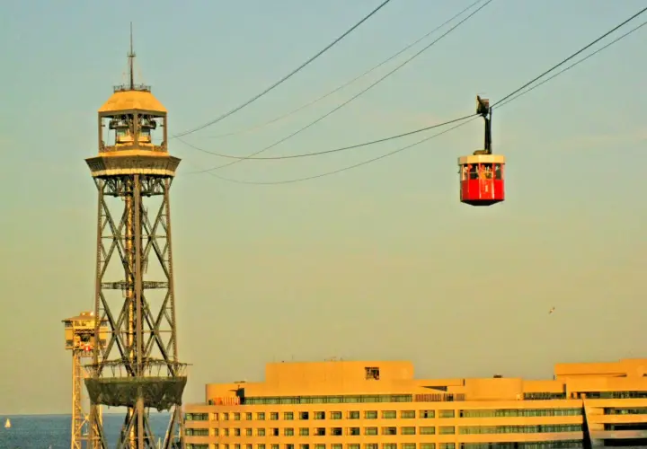 La cabina roja del montjuic seilbahn viaja sobre la ciudad, con una torre de transmisión y edificios modernos de fondo.