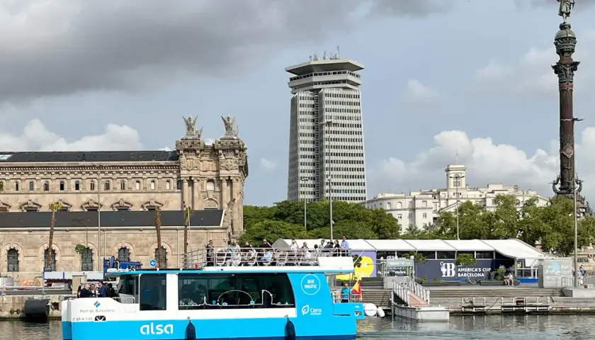 Barco turístico azul navega en el hafen Barcelona, con edificios históricos y modernos de fondo.