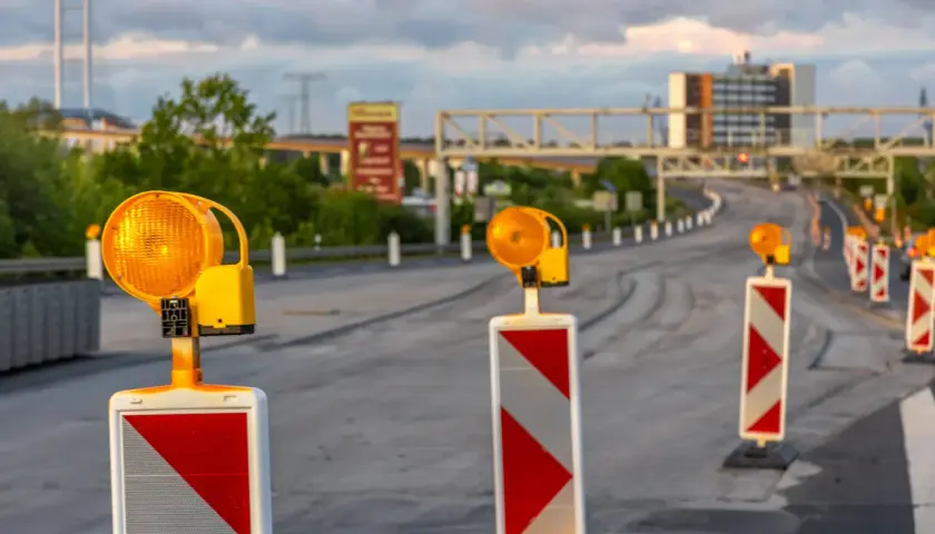 Una hilera de postes con luces naranjas intermitentes y marcas rojas y blancas, una señal de balizamiento, guía el tráfico en una carretera en construcción.
