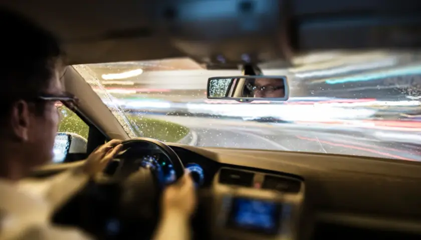Conduciendo de noche bajo la lluvia, las luces de la carretera se difuminan. El conductor, con gafas, se refleja en el retrovisor, mostrando que es el ASR de un coche.