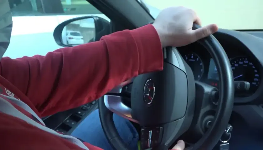 Mano girando el volante, se escucha un ruido al girar el volante. El conductor lleva una sudadera roja y va en un coche.