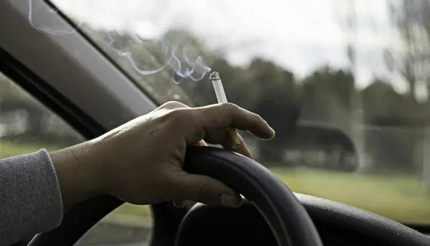 Mano sujetando un cigarrillo encendido sobre el volante. El humo se eleva mientras se puede fumar conduciendo.