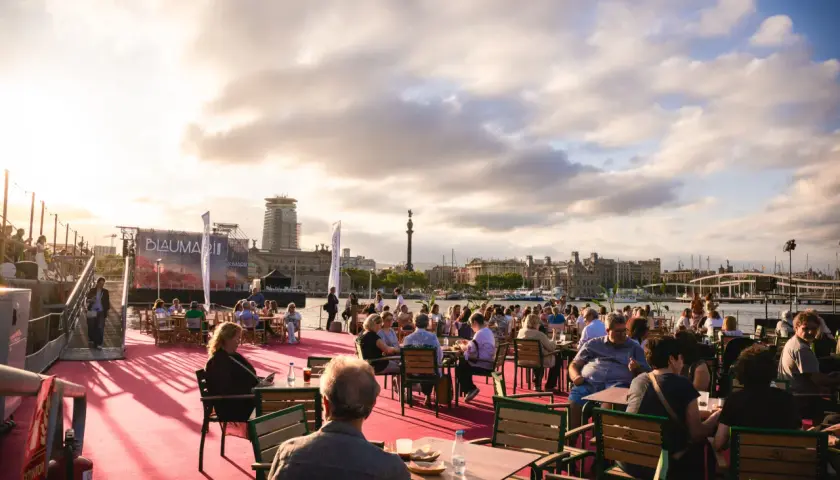 Gente disfrutando en terrazas al aire libre en el bsm moll de la fusta, con el mar y edificios de Barcelona de fondo.