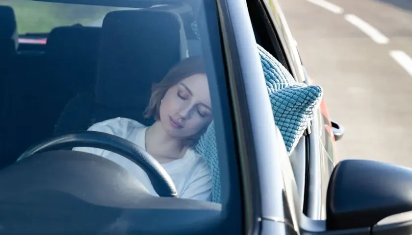 Mujer rubia duerme en el coche, apoyada en una almohada azul. Parece que se puede dormir en el coche cómodamente.
