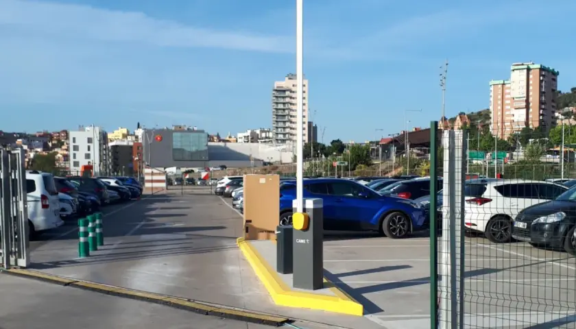 Amplio parking BSM Vall d'Hebron con coches aparcados y barrera de acceso. Edificios modernos y colinas al fondo.