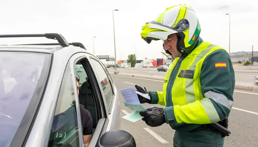 Agente de la Guardia Civil, con casco y chaleco reflectante, se acerca a un coche para entregar un documento. Parece que prescribe una multa.
