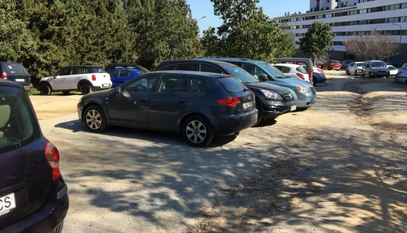 Aparcamiento del zoo de Barcelona con varios coches aparcados bajo árboles frondosos y un edificio moderno al fondo.