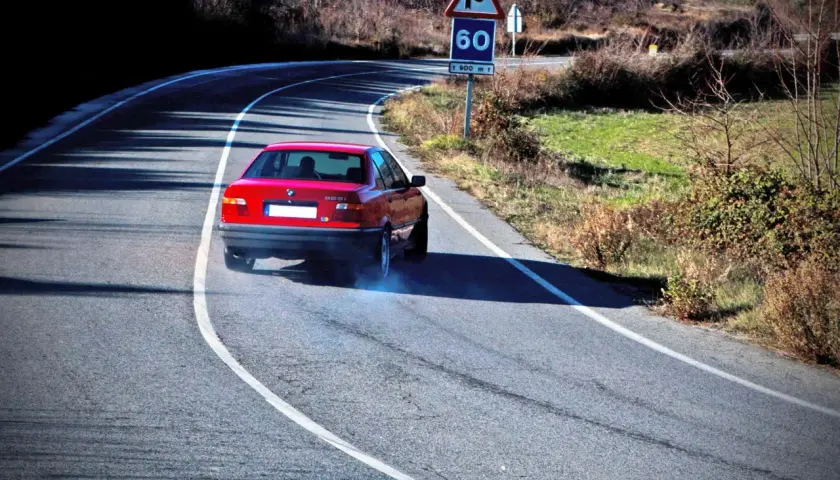 Coche rojo derrapando en curva, provocando subviraje y sobreviraje. Humo azul sale de las ruedas. Señal de curva peligrosa y límite de 60.