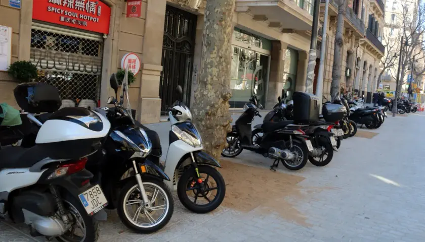 Un estacionamiento de motos T1 en una calle urbana. Varias motos y scooters están aparcados en la acera frente a edificios con tiendas y restaurantes.