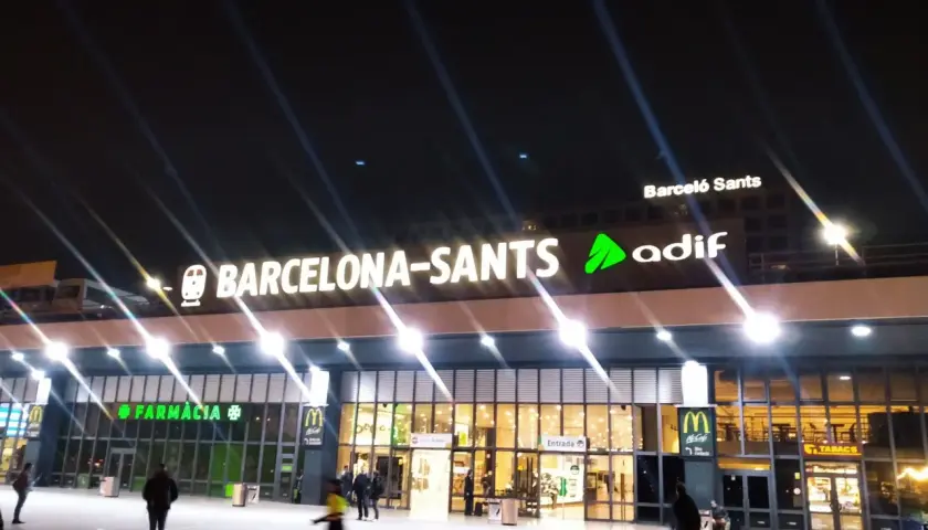 Noche en Barcelona Sants. Luces brillantes iluminan la fachada de la estación, con letreros de "Farmàcia" y "McDonald's".