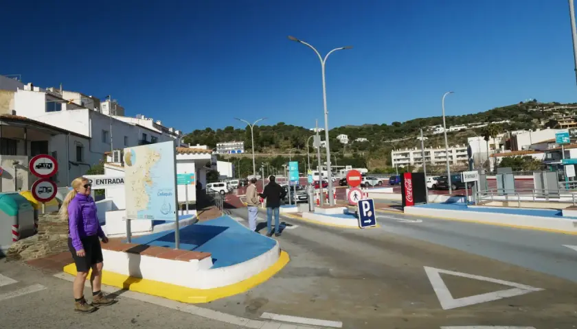 Entrada a parking gratuit Cadaqués. Una mujer rubia sonríe cerca de un mapa turístico. El cielo azul y las casas blancas dominan la escena.