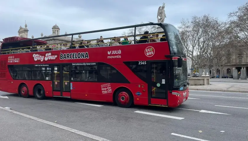 Autobús rojo de dos pisos "City Tour Barcelona" con turistas en la cubierta superior, ideal para el transporte en Barcelona para turistas.