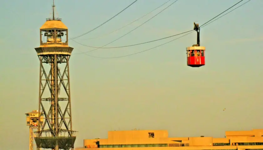 La cabina roja del montjuic seilbahn viaja sobre la ciudad, con una torre de transmisión y edificios modernos de fondo.
