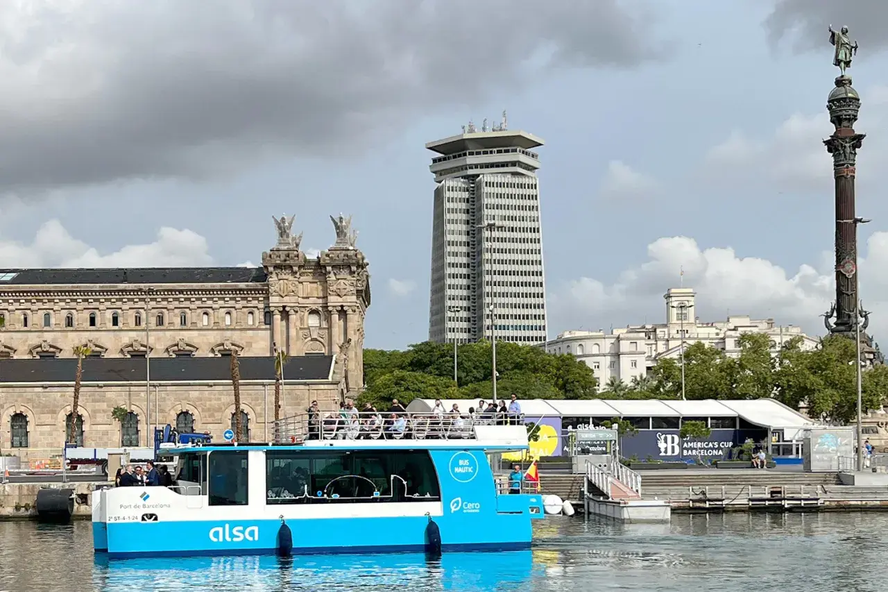 Barco turístico azul navega en el hafen Barcelona, con edificios históricos y modernos de fondo.