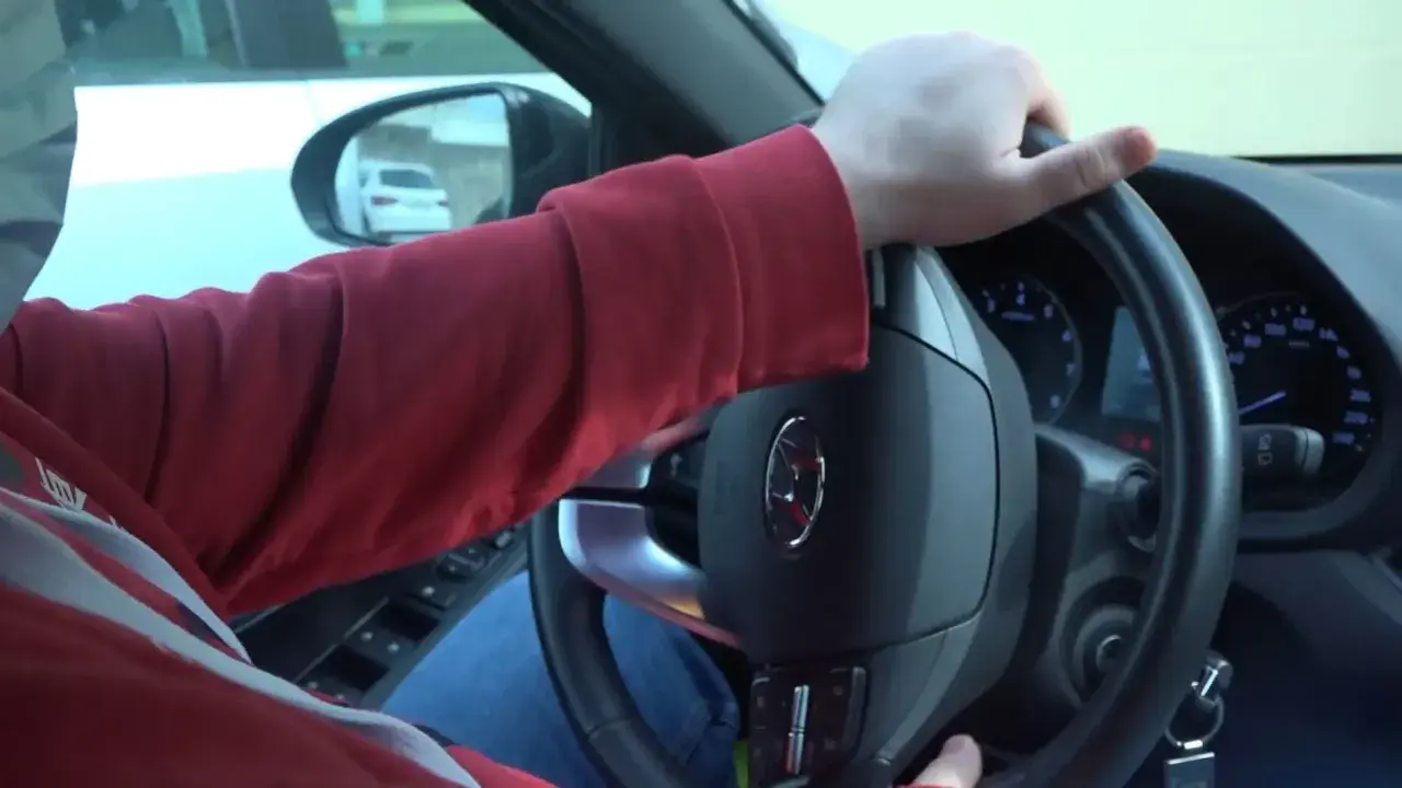 Mano girando el volante, se escucha un ruido al girar el volante. El conductor lleva una sudadera roja y va en un coche.