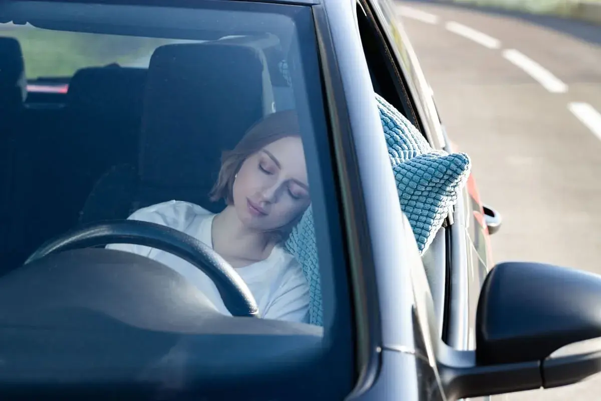 Mujer rubia duerme en el coche, apoyada en una almohada azul. Parece que se puede dormir en el coche cómodamente.
