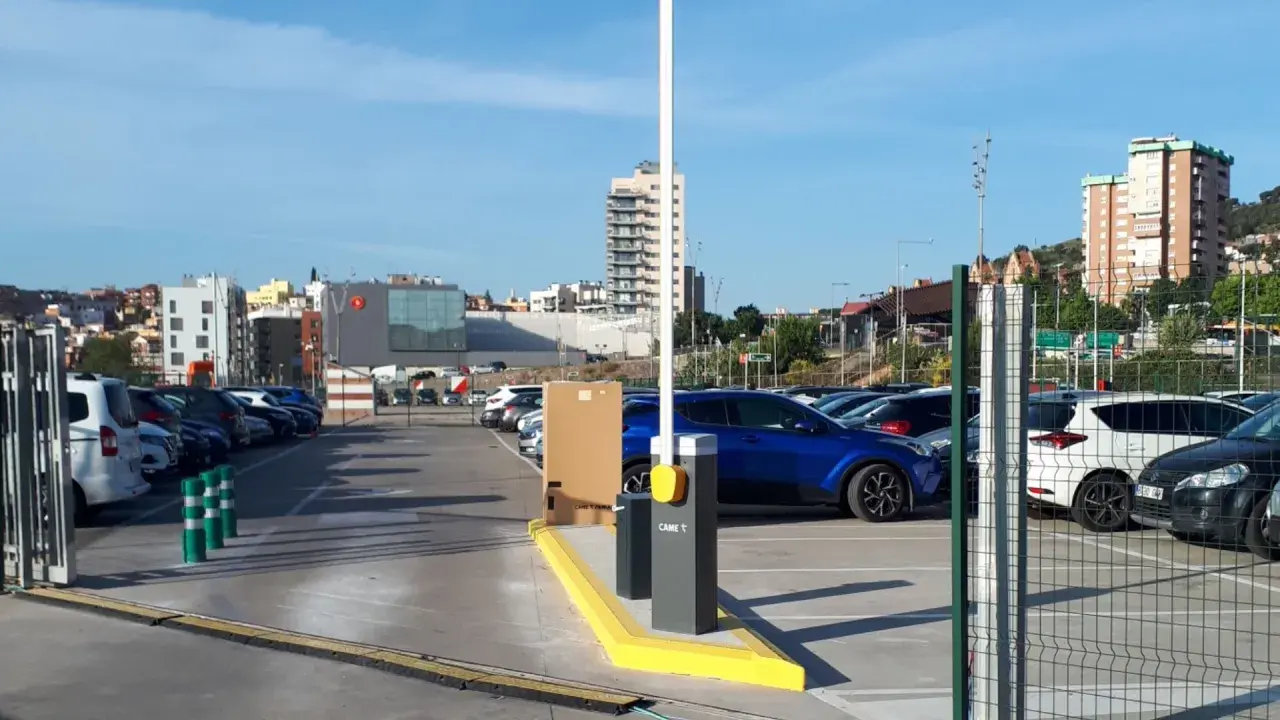 Amplio parking BSM Vall d'Hebron con coches aparcados y barrera de acceso. Edificios modernos y colinas al fondo.