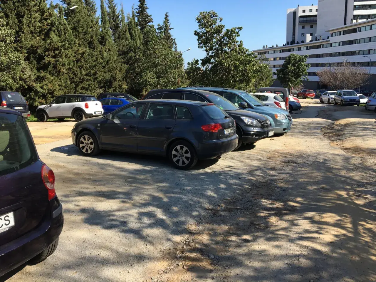 Aparcamiento del zoo de Barcelona con varios coches aparcados bajo árboles frondosos y un edificio moderno al fondo.