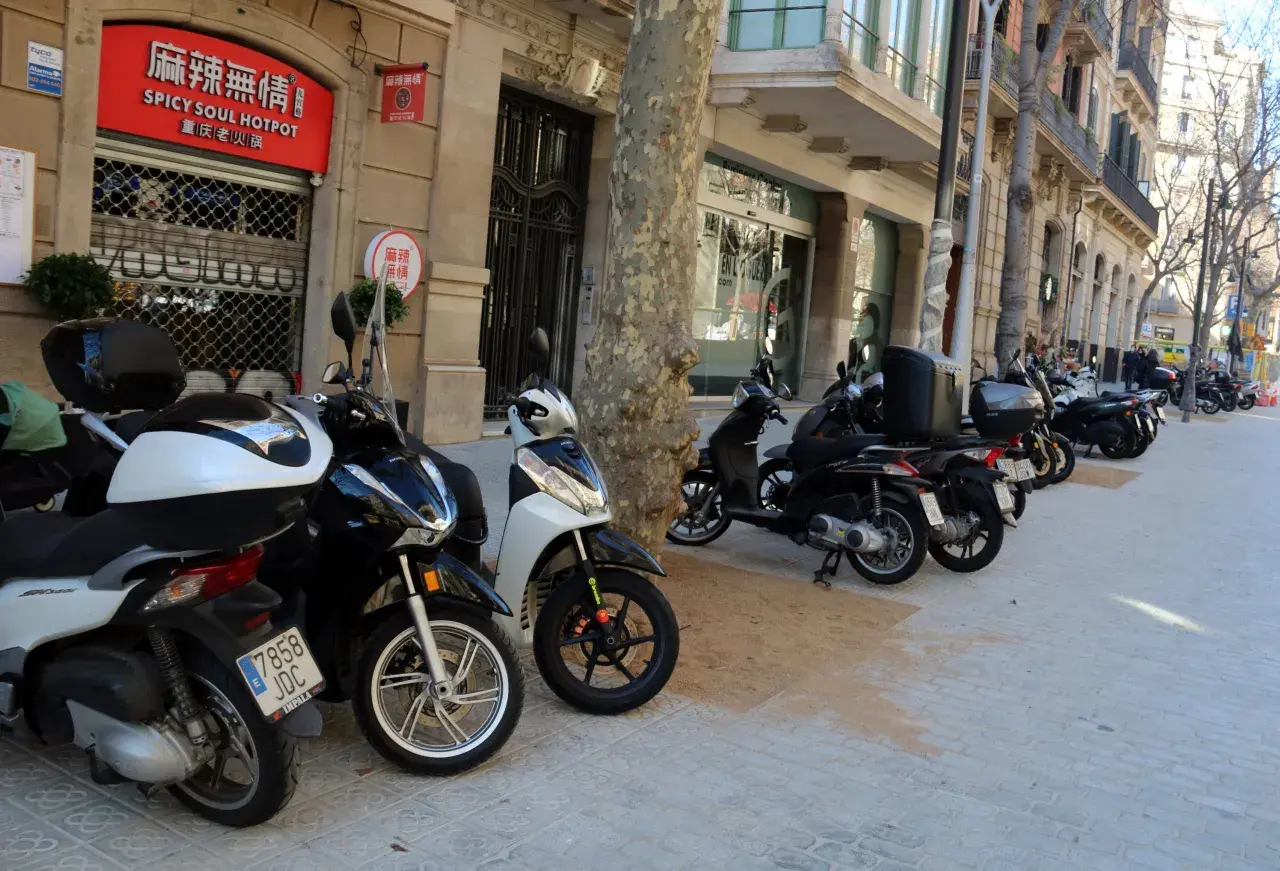 Un estacionamiento de motos T1 en una calle urbana. Varias motos y scooters están aparcados en la acera frente a edificios con tiendas y restaurantes.